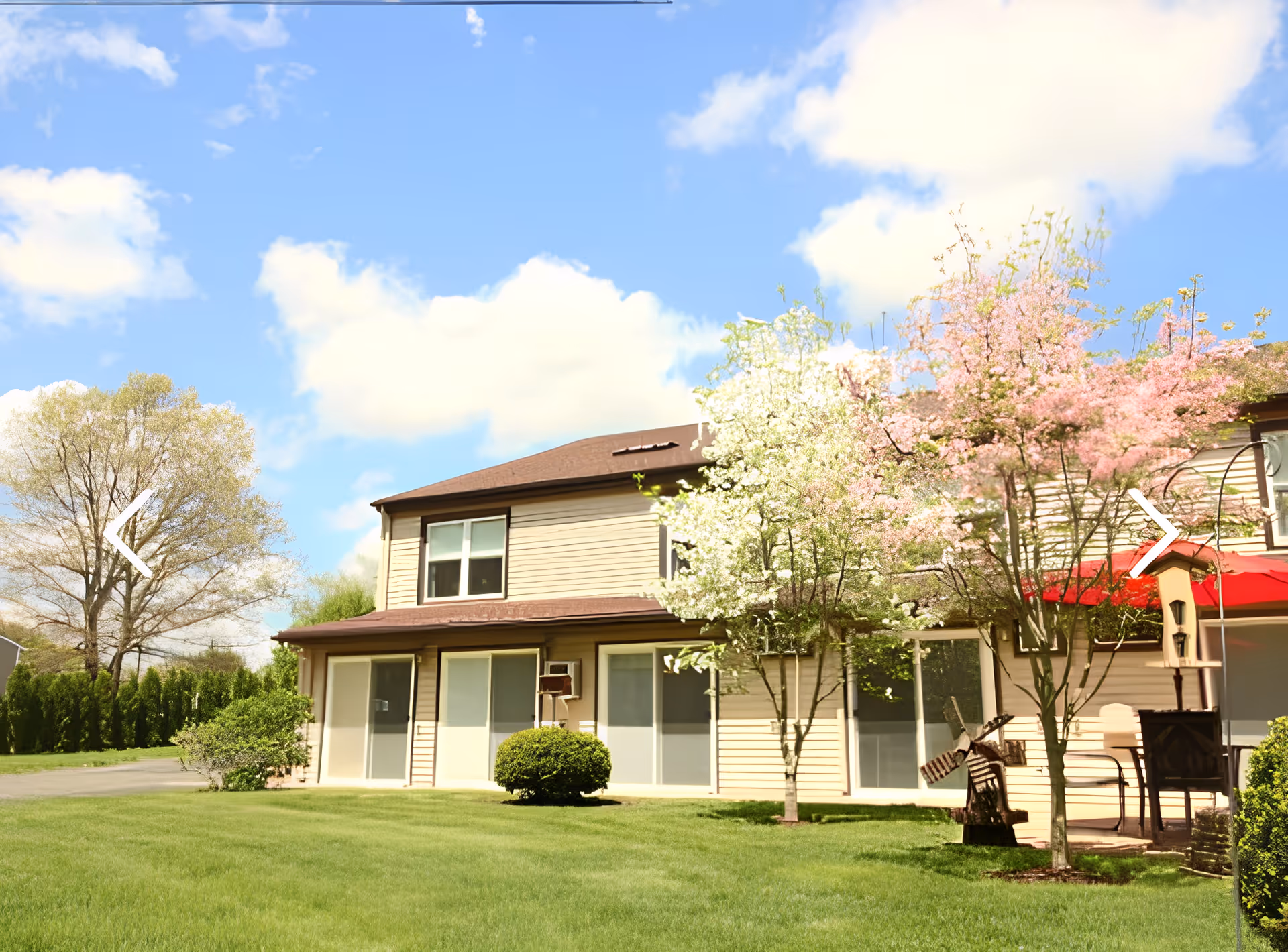 Front exterior of a two-story residential building with a green lawn, blooming trees, and a small patio area.