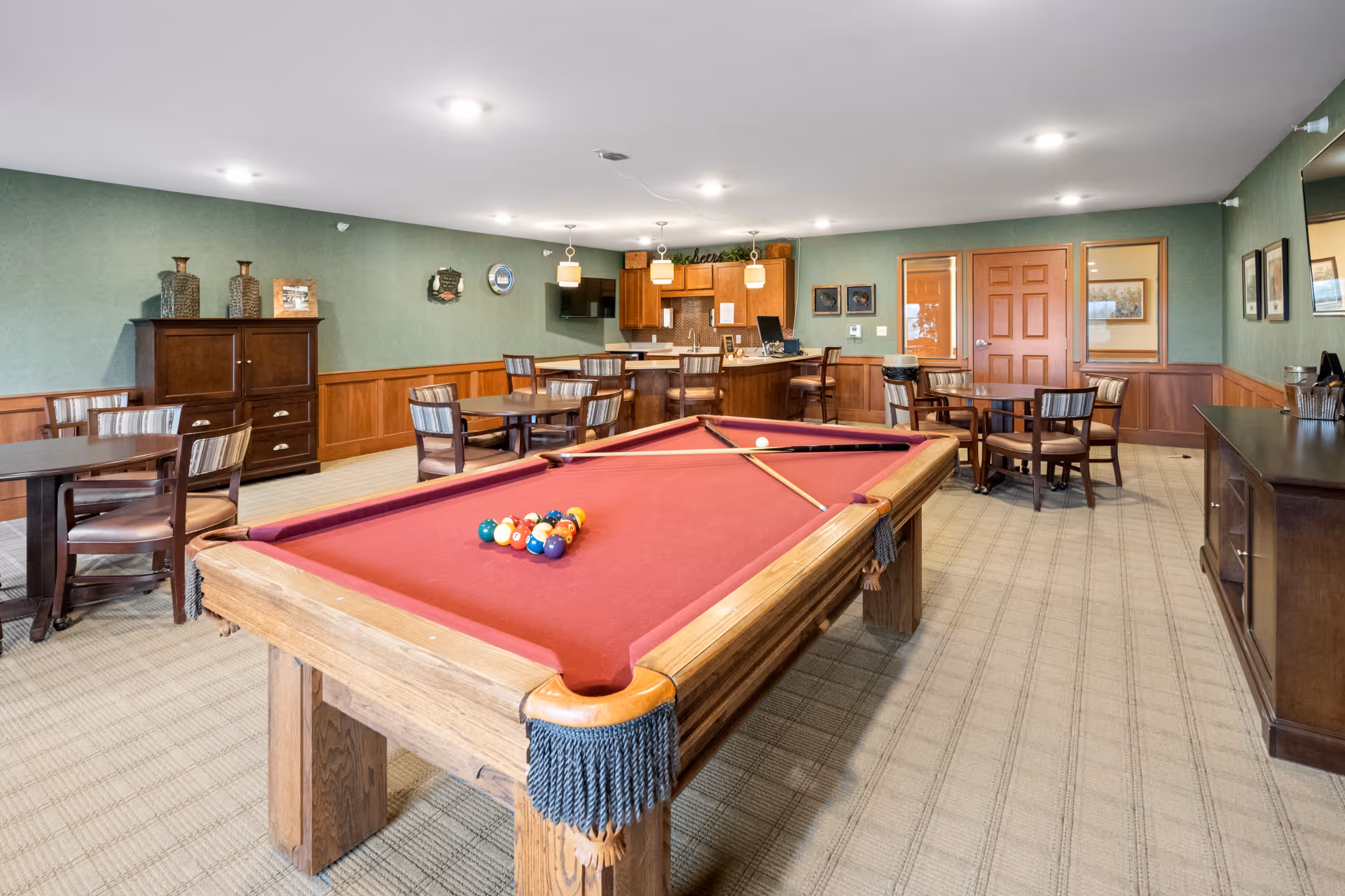 A spacious recreational room with a red felt pool table in the center, surrounded by several wooden tables and chairs. The room has green walls with wood paneling, a carpeted floor, and a small kitchen area with bar stools in the background. There are decorative items on the walls and furniture, and the room is well-lit with ceiling lights.