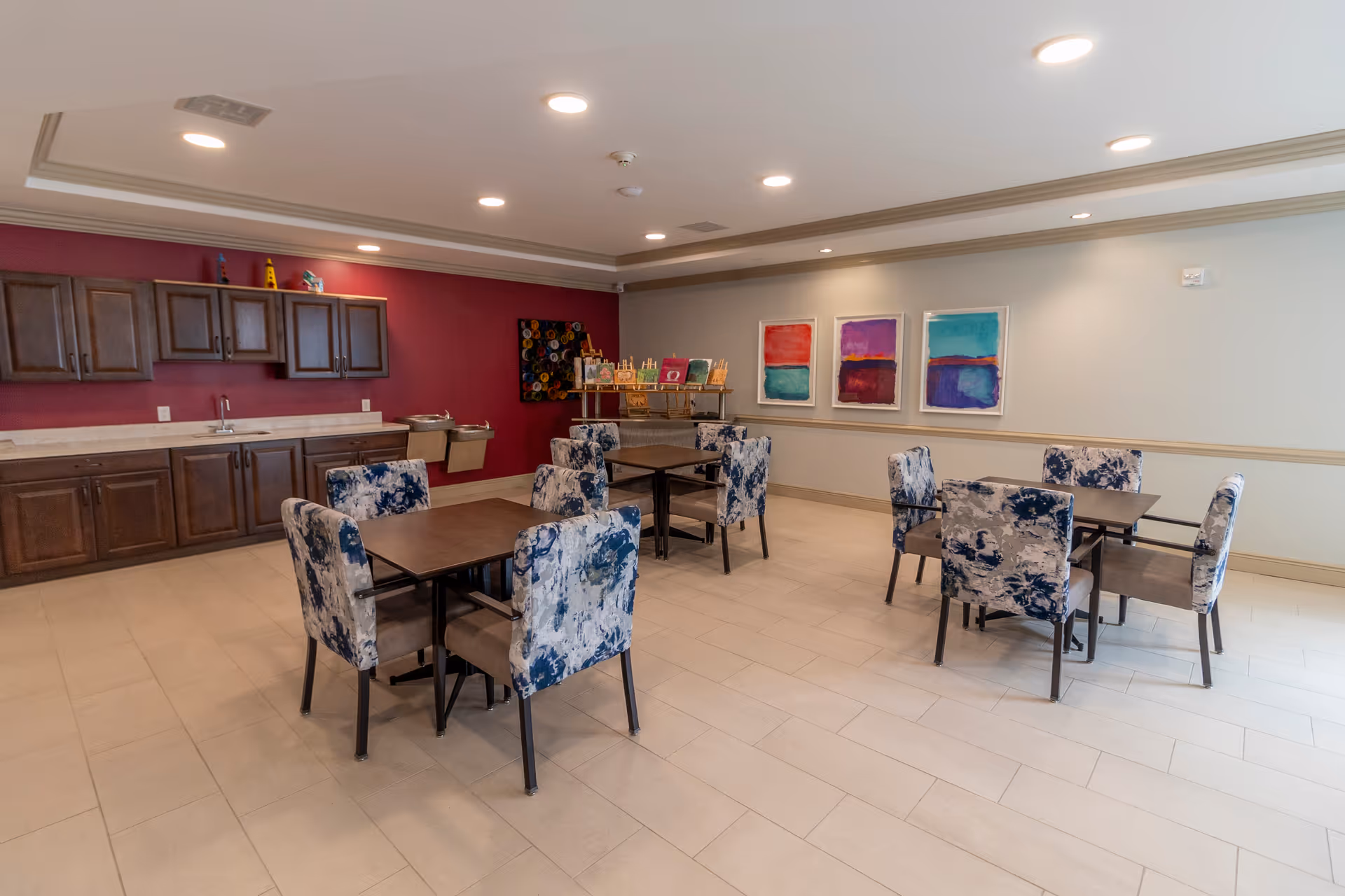 Well-lit communal dining room with several square tables and patterned chairs, a kitchenette along a red wall, and colorful artwork on the far wall.