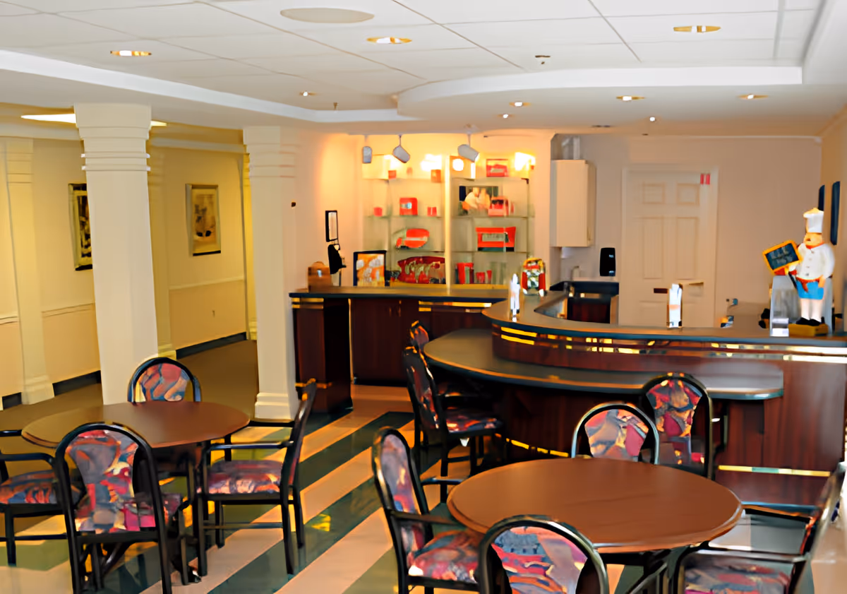 Interior view of a senior living facility dining area with round tables and chairs featuring colorful upholstery. A curved counter or bar area is visible in the background with decorative items and lighting. The space has a tiled floor with a striped pattern and white walls with columns.