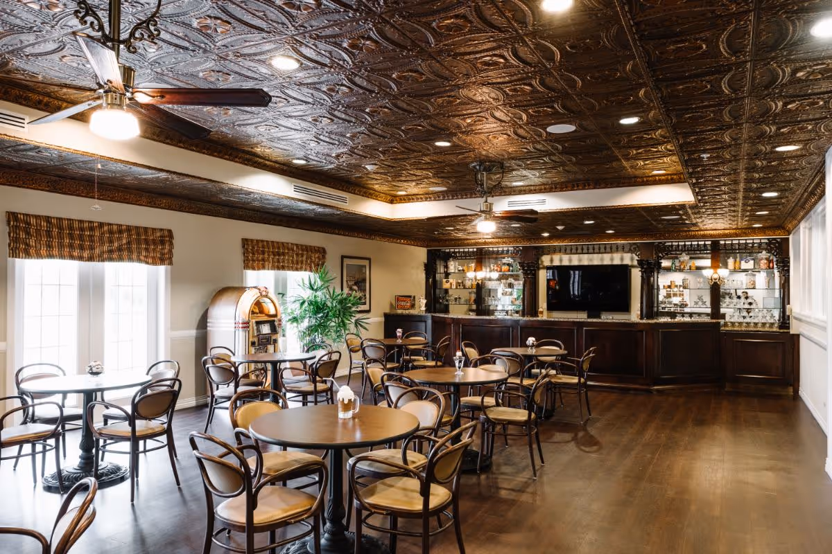 A spacious dining area with multiple round tables and wooden chairs arranged neatly. The room features a decorative, embossed tin ceiling with ceiling fans and recessed lighting. There is a bar area with glass shelves displaying various items and a large TV mounted on the wall behind the bar. Large windows with patterned valances allow natural light to fill the room, and a jukebox and a potted plant are visible near the windows.