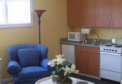 A small living area with a blue upholstered armchair next to a floor lamp, adjacent to a kitchenette with wooden cabinets, a microwave, a small refrigerator, a sink, and a stove. A round table in the foreground holds a floral arrangement.