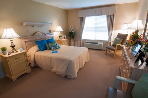 Sunny bedroom with a bed, bedside tables and lamps, dresser and an armchair by a window.