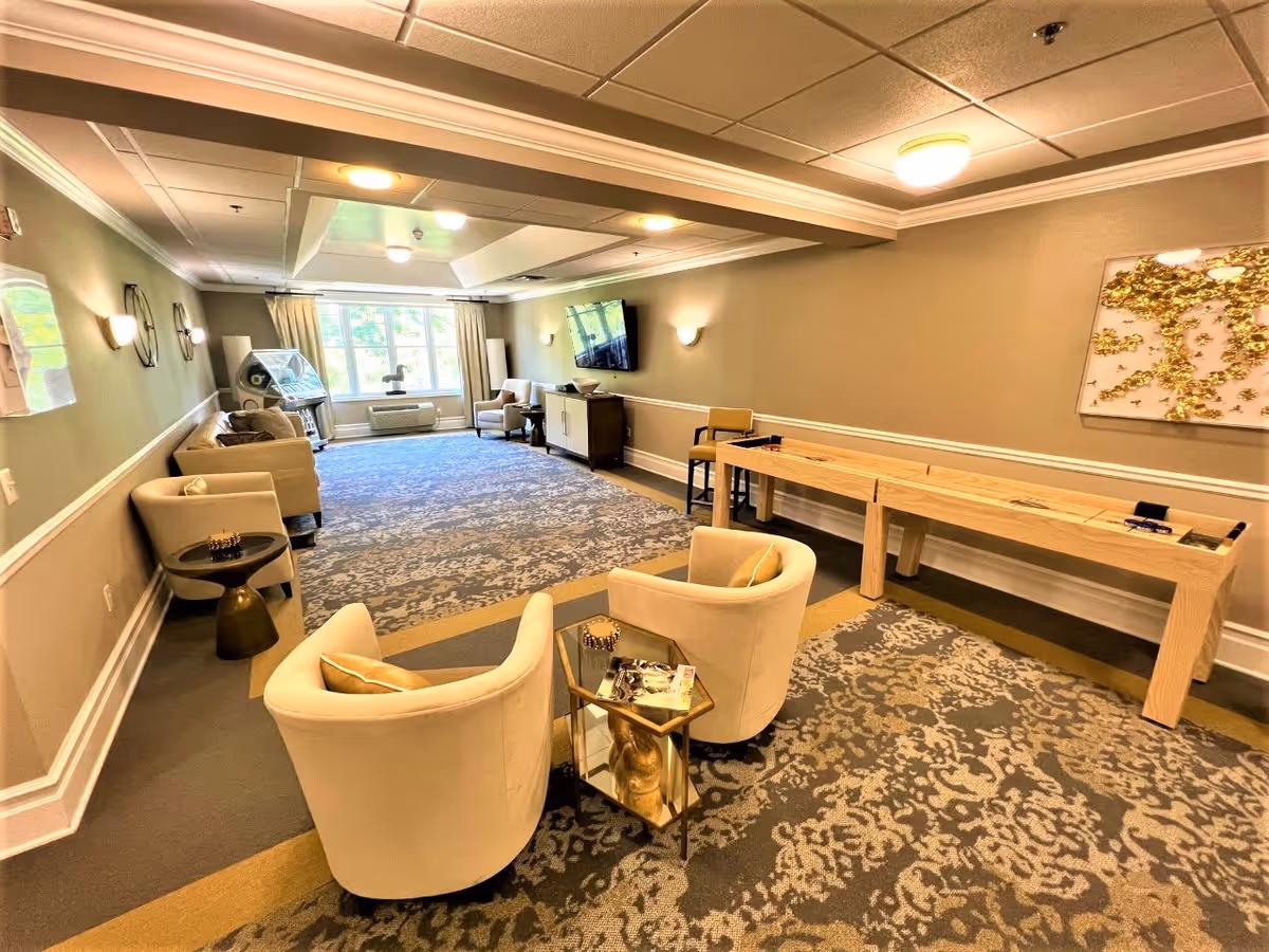 A spacious, well-lit lounge area with beige walls and patterned carpet. The room features several cream-colored armchairs arranged around small tables, a long wooden shuffleboard table along one wall, a wall-mounted flat-screen TV, and a large window with curtains letting in natural light. Decorative wall art and sconces add to the cozy ambiance.