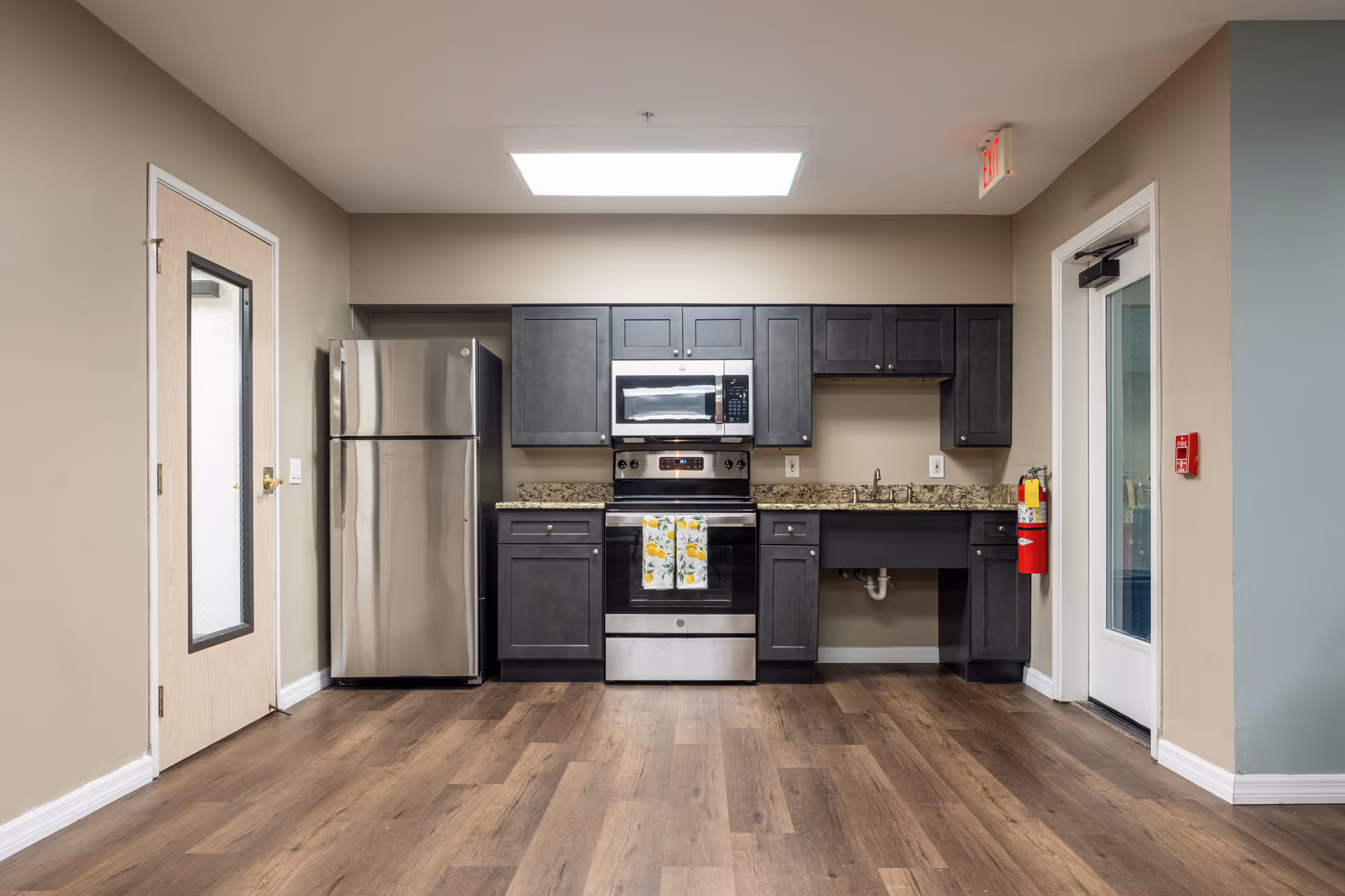 A modern kitchen area with dark gray cabinets, a stainless steel refrigerator, stove, and microwave. The countertop is granite, and there is a fire extinguisher mounted on the wall near a glass door. The floor is wood, and there is a skylight above providing natural light.
