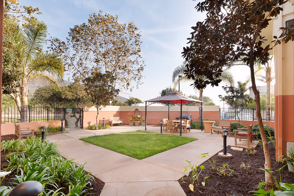 Sunny landscaped courtyard with a central lawn, gazebo seating, benches and surrounding plants at a senior living facility.