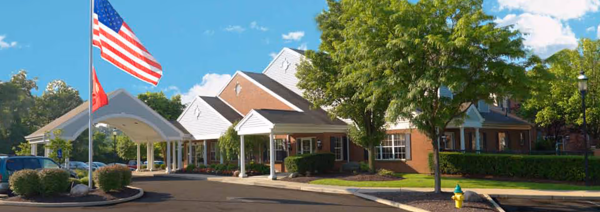 Front entrance of a brick senior living facility with a covered drive, flagpoles, trees, and a parking area.