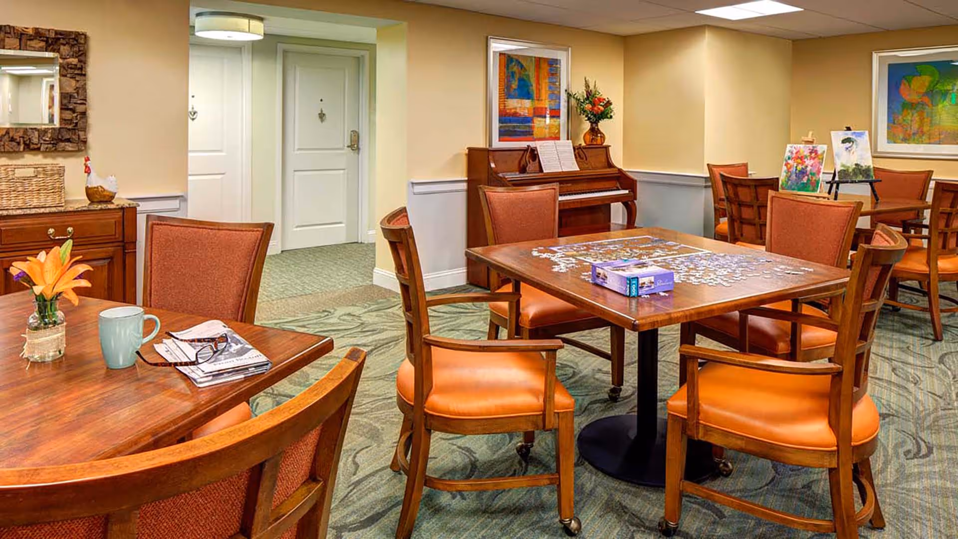 A cozy common room with wooden tables and orange cushioned chairs. One table has a partially completed jigsaw puzzle and a puzzle box on it. Another table has a vase with an orange flower, a coffee mug, a pair of glasses, and some newspapers. In the background, there is an upright piano with sheet music and a vase of flowers on top. The walls are decorated with colorful framed artwork, and there are easels displaying paintings. The room has a carpeted floor with a patterned design and soft yellow walls.