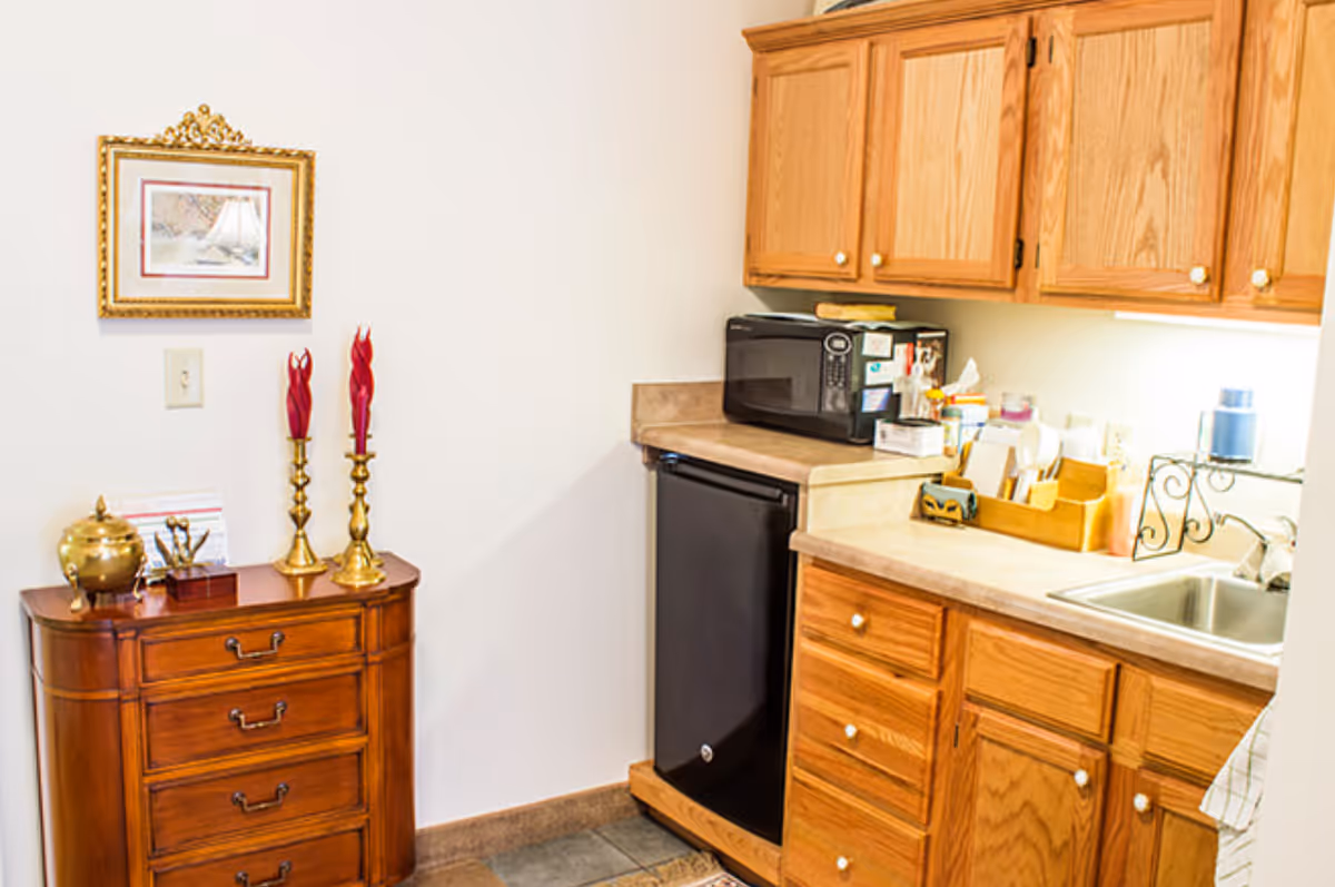 A small kitchen area with wooden cabinets, a black mini refrigerator, a microwave on the countertop, a stainless steel sink, and various kitchen items. To the left, there is a wooden chest of drawers with decorative items including two red candles in brass holders and a framed picture on the wall above.