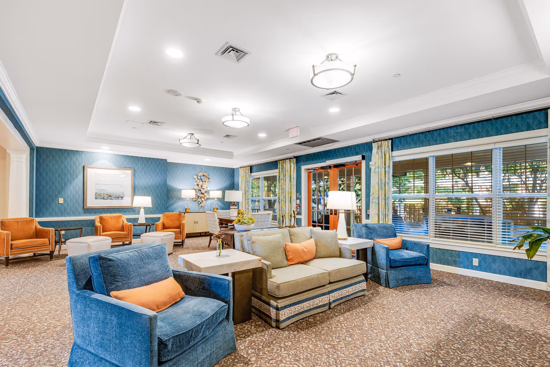 A bright and spacious living room area in a senior living facility with blue and orange upholstered chairs, a beige sofa with orange pillows, side tables with lamps, large windows with blinds, and blue patterned wallpaper. The room has a carpeted floor and ceiling lights, with a door leading outside.