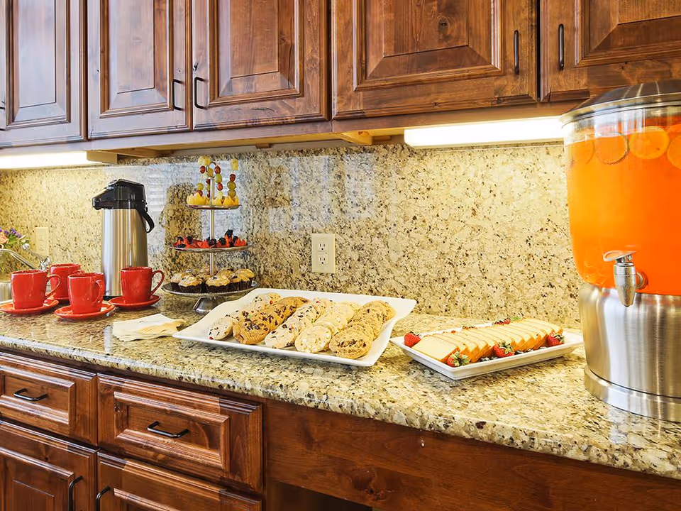 A kitchen counter with a granite countertop and wooden cabinets above and below. On the counter, there are red coffee cups with saucers, a stainless steel coffee dispenser, a tiered tray with muffins and fruit skewers, a plate of assorted cookies, a plate of sliced cheese with strawberries, and a large beverage dispenser filled with orange drink and slices of citrus fruit.