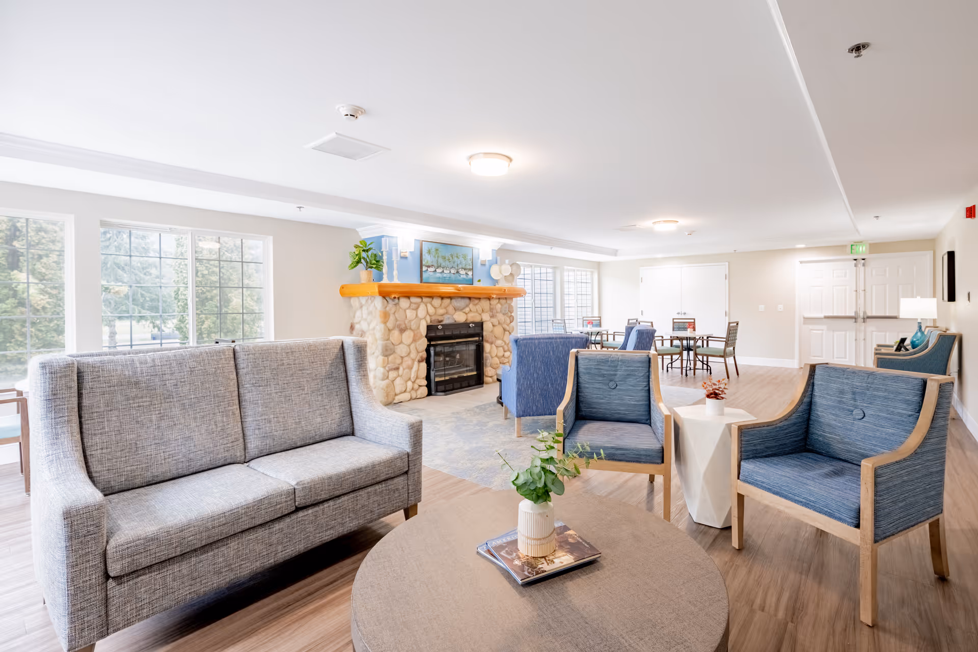 A bright and spacious living room area in Birchview Memory Care featuring a gray sofa, two blue armchairs, a round wooden coffee table with a small plant and book, a stone fireplace with a wooden mantle, large windows letting in natural light, and several tables and chairs in the background.