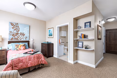 A cozy bedroom in a senior living facility featuring a bed with a coral-colored bedspread and decorative pillows, a nightstand with a lamp, a chest of drawers, and an open doorway leading to a bathroom. The room has beige walls, carpeted floor, and built-in wall shelves with framed pictures and decorative items.