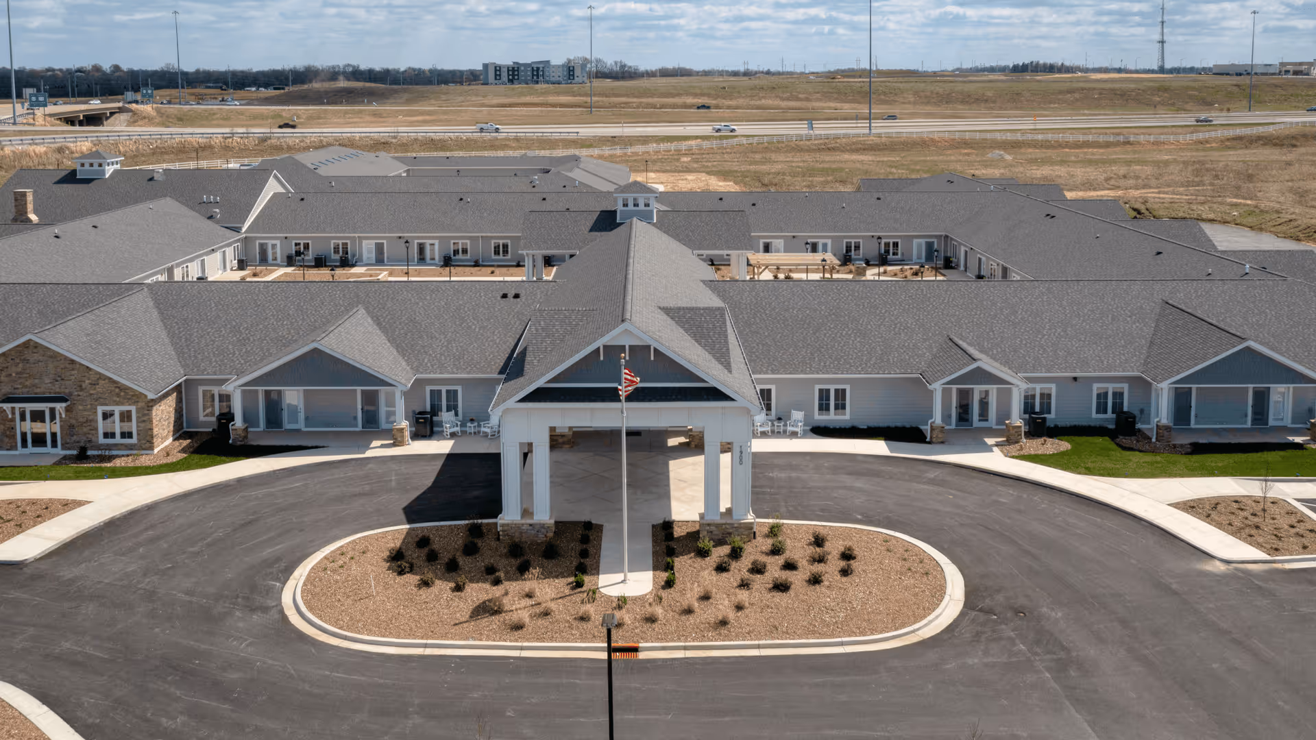 Aerial view of Cedarhurst Senior Living of Owensboro building with a large covered entrance, circular driveway, and landscaped area in front. The building has a gray roof and light-colored exterior walls, situated in an open area with a highway visible in the background.