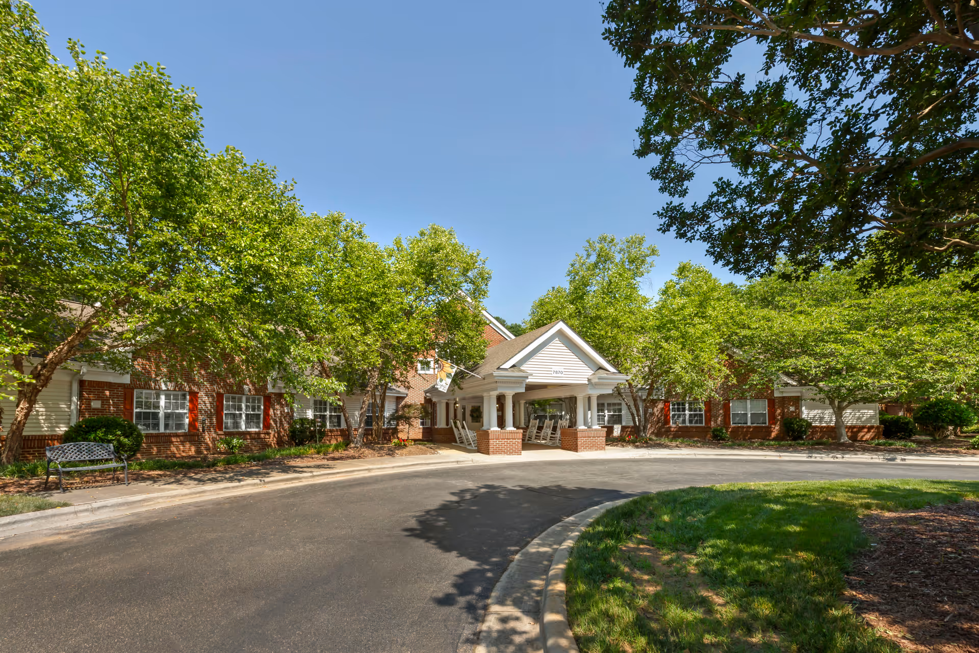 Exterior view of Brookdale Cary senior living facility with a circular driveway, brick building, white entrance canopy supported by columns, surrounded by green trees and clear blue sky.