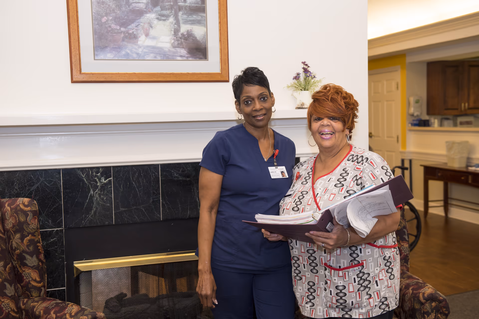 Two women standing indoors in front of a fireplace with a framed picture above it. One woman is wearing navy blue scrubs and the other is wearing patterned scrubs holding an open binder with papers. They are smiling and appear to be staff members in a senior living facility.