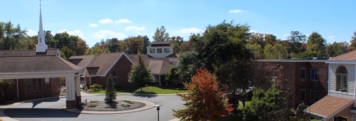 Exterior view of Wesbury United Methodist facility showing multiple connected brick buildings with brown roofs, a white steeple, a circular driveway with landscaped greenery, and trees with autumn foliage under a clear blue sky.