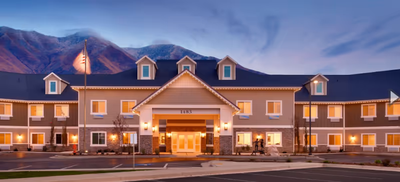 Front view of Spring Gardens Senior Living Mapleton exterior with lit windows, mountains, and parking.