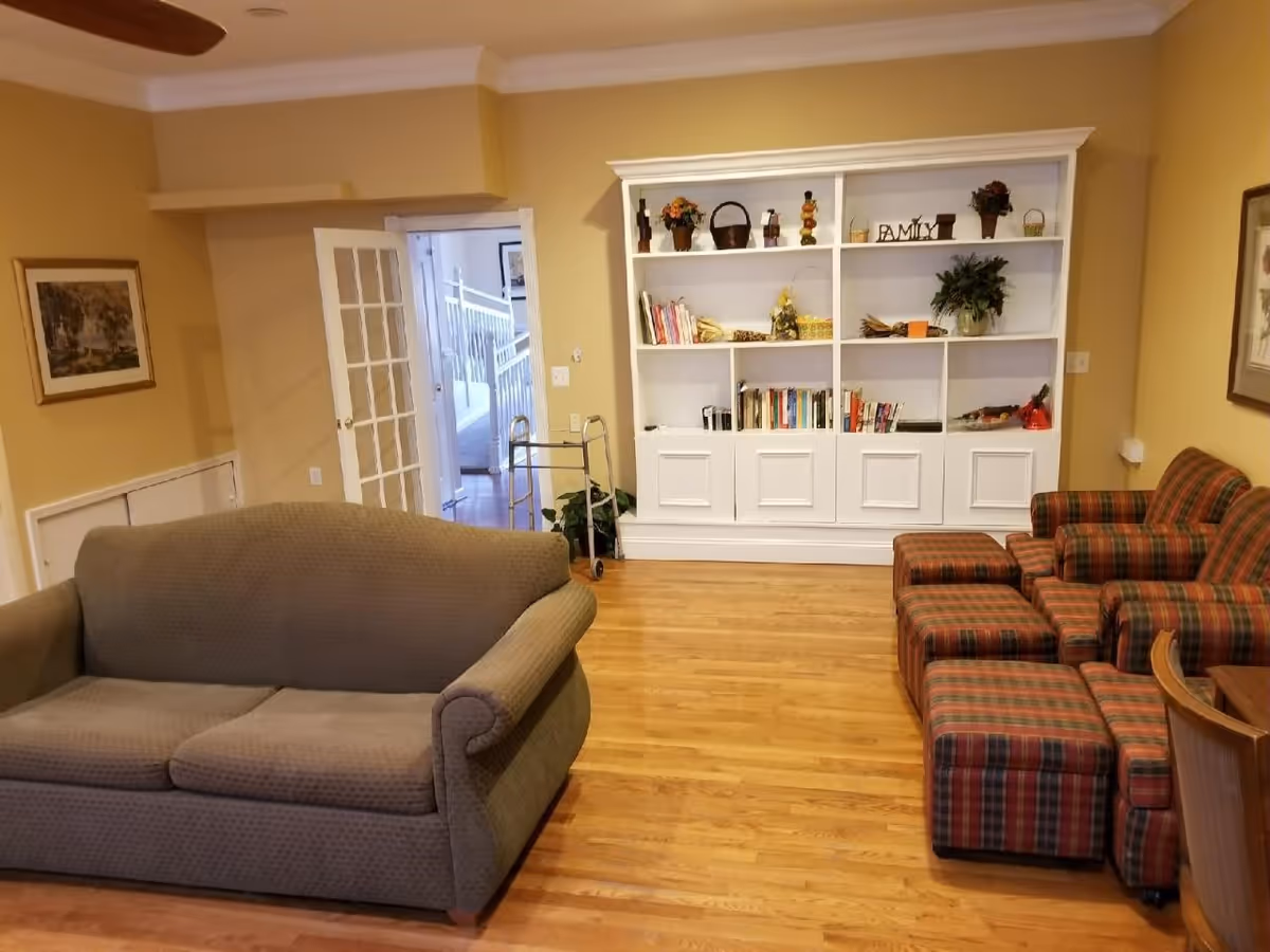 Well-lit living room with a gray sofa, plaid armchairs and ottomans, a white built-in bookshelf, and hardwood floors.