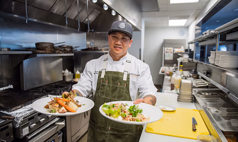 A chef wearing a white coat, green apron, and black cap stands in a commercial kitchen holding two plates of food, one with a dish featuring vegetables and the other with a salad. The kitchen has stainless steel appliances, cooking utensils, and a yellow cutting board with a knife on the counter.