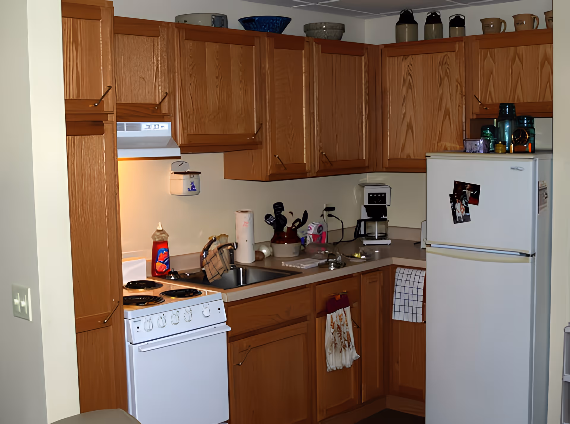 Small kitchen with wooden cabinets, a white stove and refrigerator, sink, coffee maker and various items on the countertops.