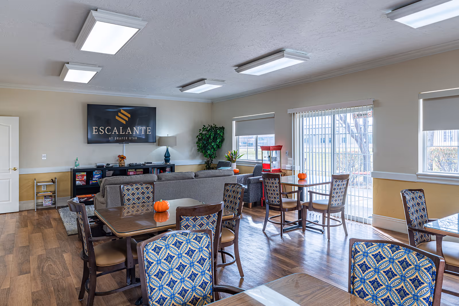 Communal dining and lounge area with tables and patterned chairs, sofas, a TV reading "Escalante", and small pumpkins on the tables.