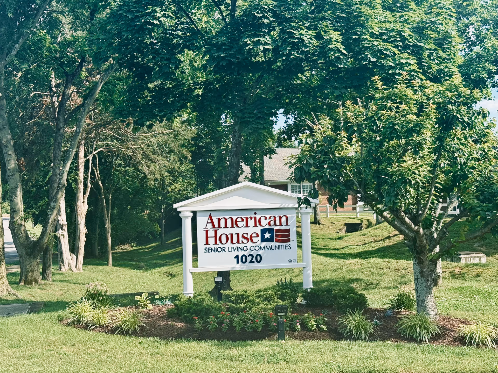 White sign reading "American House Senior Living Communities 1020" on a landscaped lawn with trees and a building in the background.