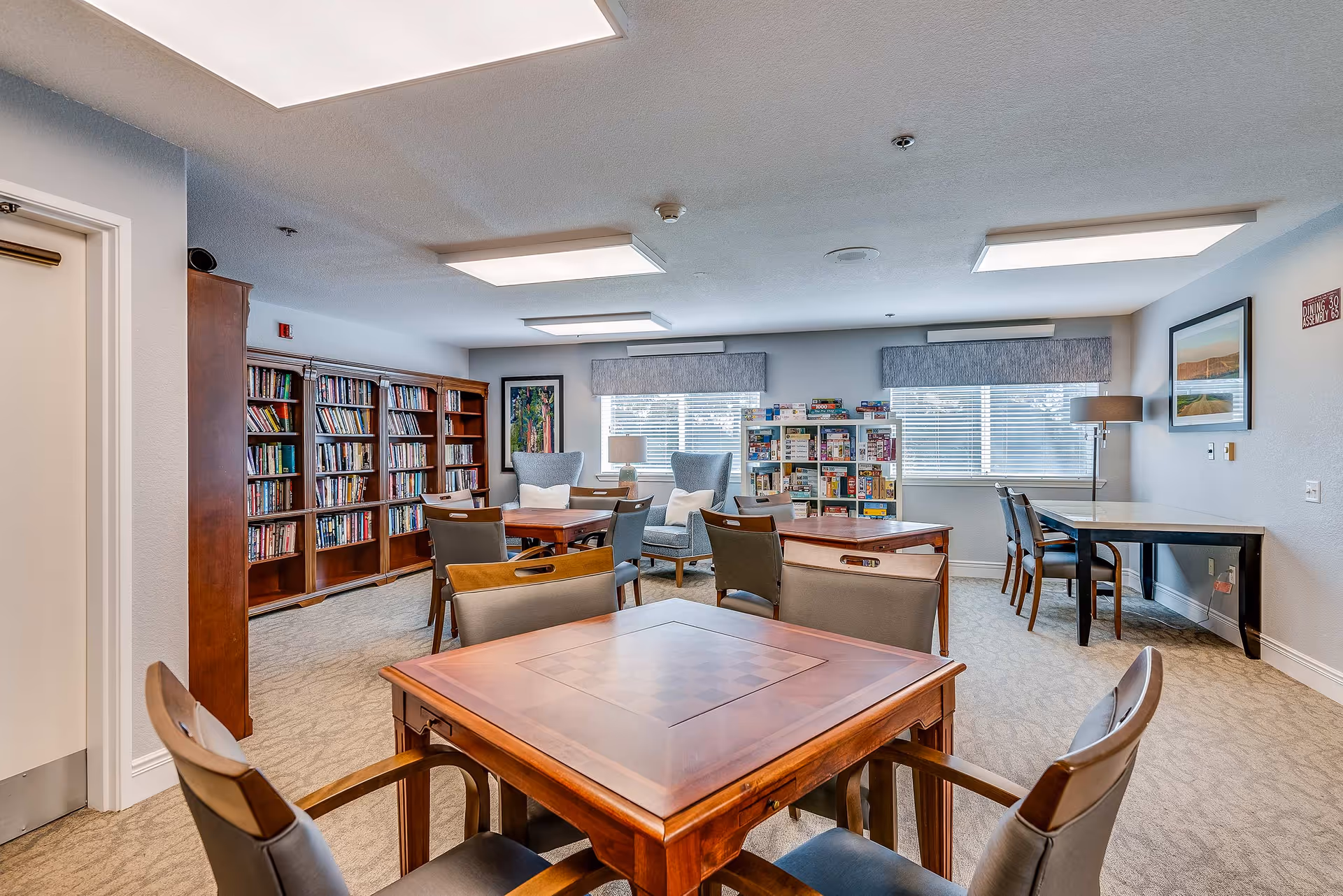 A bright senior common room with multiple game tables, bookshelves, chairs, and windows.