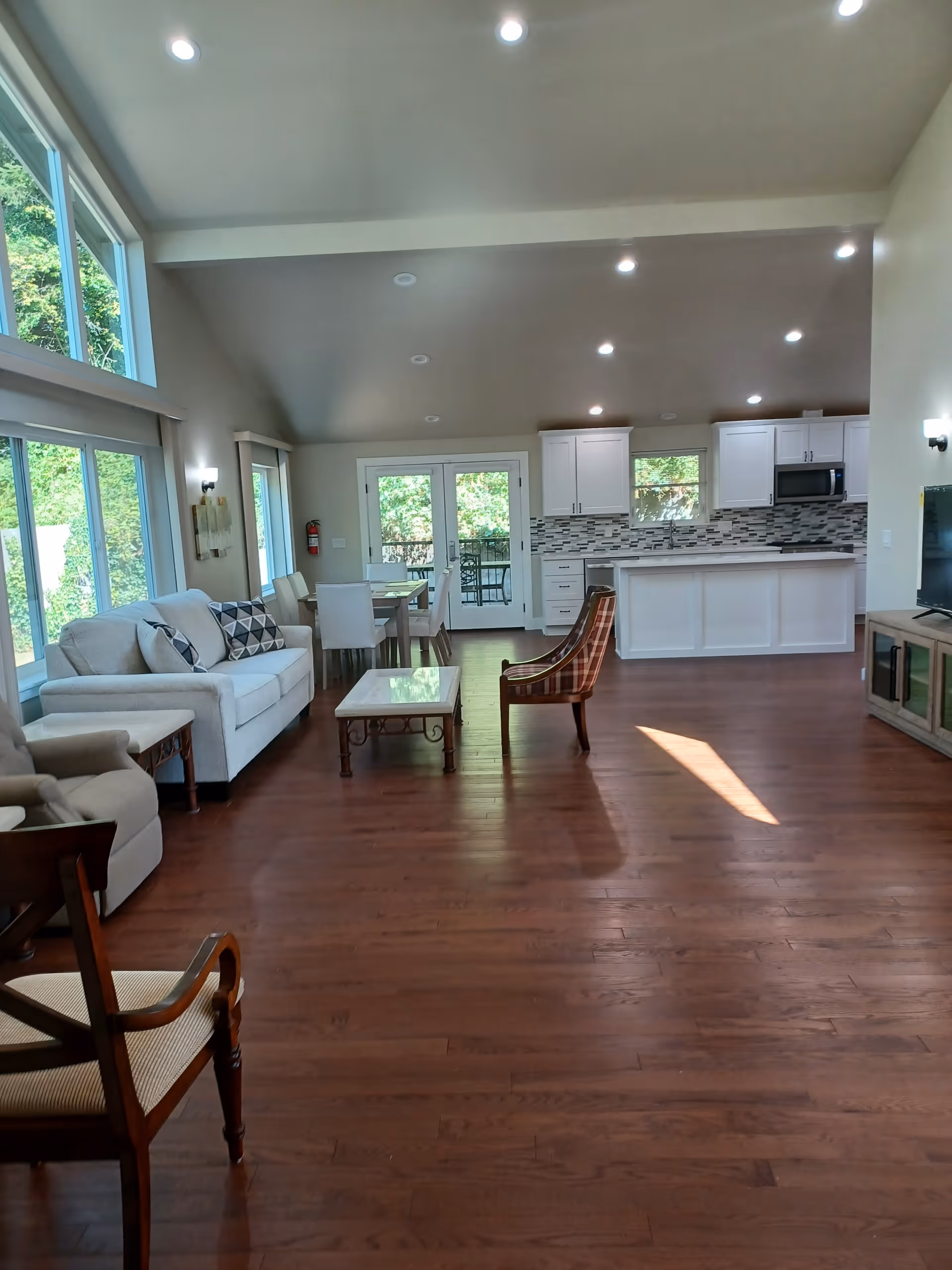 Spacious open-plan living room and kitchen with hardwood floors, large windows, white cabinets, and seating.