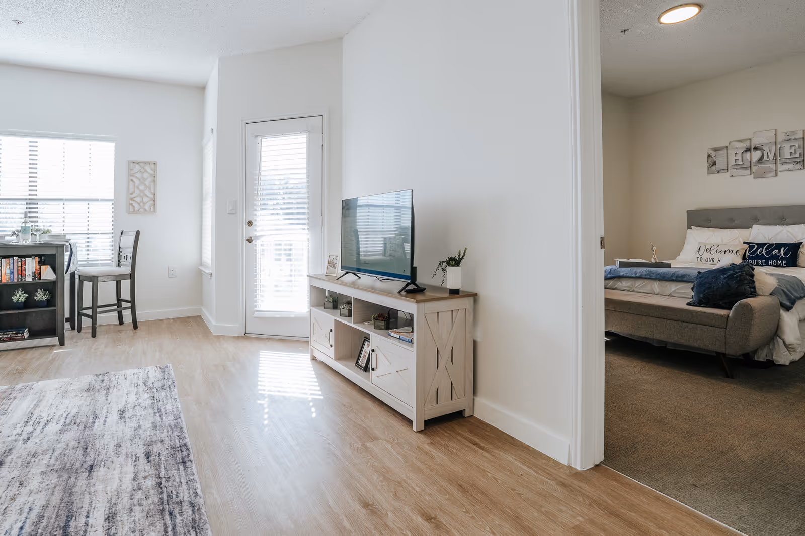 A bright and airy living space with light wood flooring, a TV on a wooden stand, a small dining table with two chairs near a window, and a doorway leading to a bedroom with a bed, pillows, and wall art.