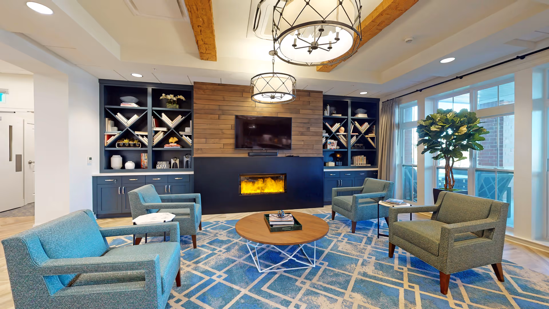 A cozy living room area with four teal armchairs arranged around a round wooden coffee table on a blue patterned rug. The back wall features built-in dark blue shelves with books and decorative items, a wooden panel with a mounted flat-screen TV, and a modern electric fireplace below. Large windows on the right side let in natural light, and a tall green plant is placed near the windows. Two ceiling light fixtures with metal frames hang above the seating area.