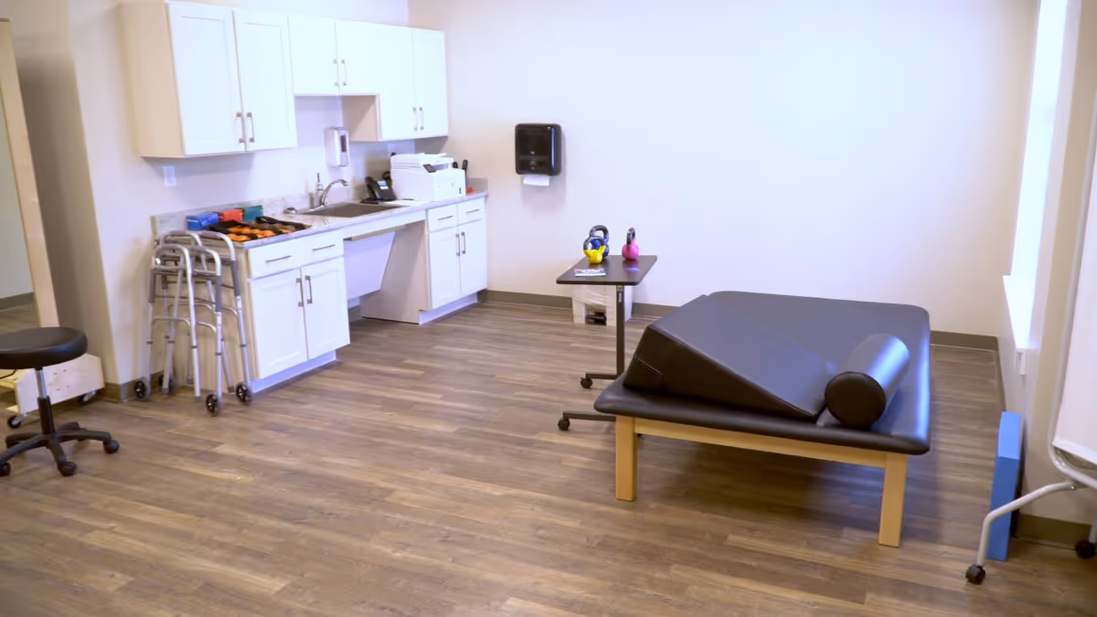 A bright therapy room with a treatment table, exercise wedges and rollers, kettlebells on a small table, cabinets and walkers on wood-look flooring.