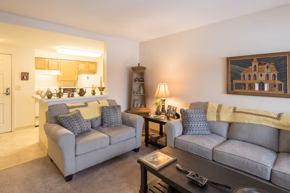 A cozy living room with two gray sofas adorned with patterned pillows and yellow throws. A wooden coffee table with decorative items sits in front of the sofas. To the right, a side table with a lamp, framed photos, and a small plant is positioned next to a corner display cabinet with shelves holding various items. The wall features a framed artwork of a house. In the background, a kitchen area with wooden cabinets, a white refrigerator, and a stove is visible.