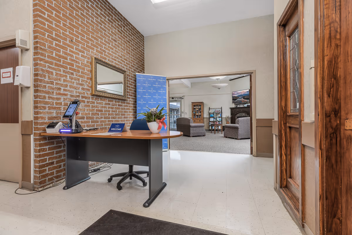 Reception desk with a welcome sign and potted plant in an entryway leading to a seating area in a senior living facility.