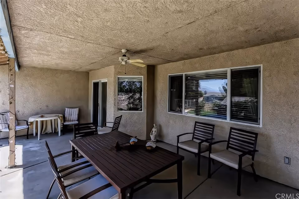 Covered outdoor patio area with a ceiling fan, a rectangular wooden table surrounded by chairs, and a small round table with two chairs in the corner. The walls are textured and there are windows and a sliding glass door visible.