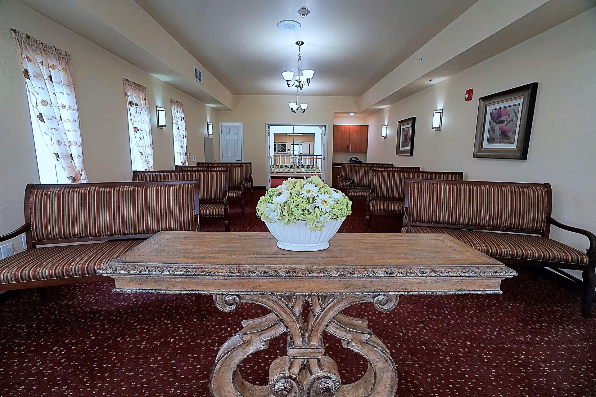 Interior view of a sitting area in a retirement living facility with striped upholstered benches arranged along the walls, a wooden table with a floral centerpiece in the foreground, framed floral artwork on the walls, and windows with sheer floral curtains letting in natural light.