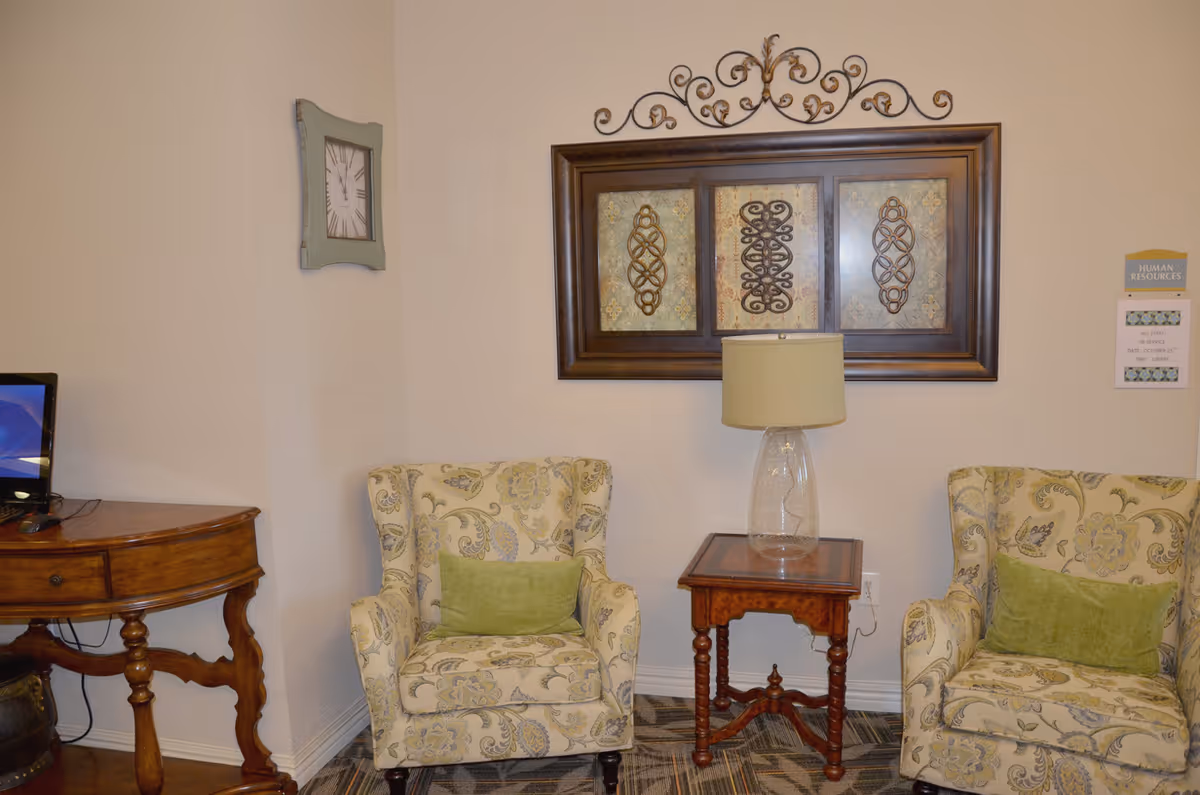 A cozy sitting area with two patterned armchairs, each with a green cushion, separated by a wooden side table with a glass lamp on it. Above the table hangs a decorative framed artwork with intricate designs. To the left, there is a wooden desk with a computer monitor. A wall clock is mounted on the adjacent wall, and a small sign labeled 'Human Resources' is visible on the right wall.