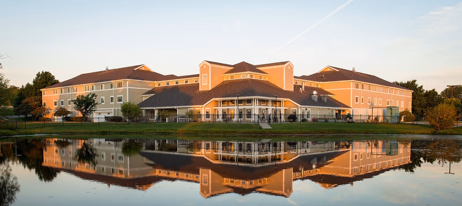 A large, multi-story building of Fort Scott Presbyterian Village reflected in a calm pond in front of it, surrounded by green grass and trees under a clear sky during sunset.