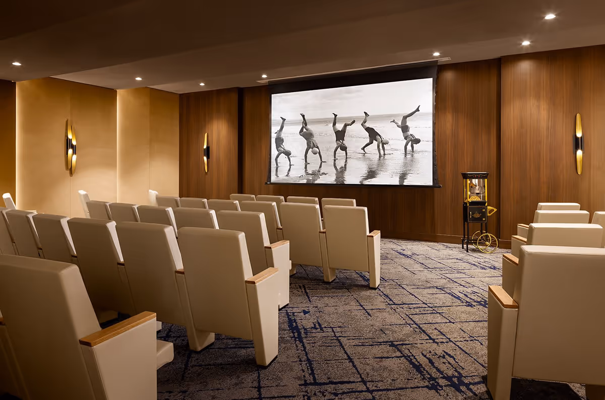 A small theater room with rows of beige cushioned chairs facing a large screen displaying a black and white photo of five people doing handstands on a beach. The room has wood-paneled walls, modern wall sconces, and a popcorn machine on the right side.