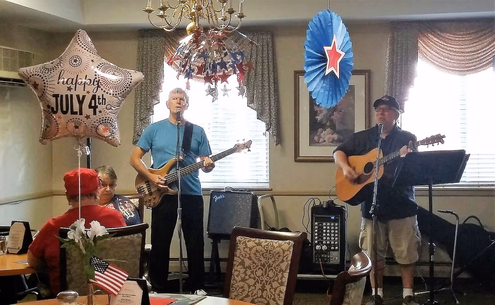 Two musicians play guitars in a decorated common dining room with seated residents and a 'Happy July 4th' balloon.
