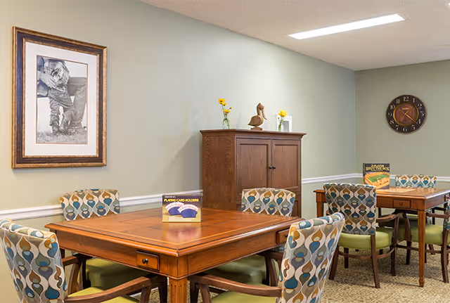 A room with two wooden tables surrounded by patterned chairs with green cushions. On the tables are boxes labeled 'Playing Card Holders' and 'Scrabble Super Crossword'. A wooden cabinet with decorative items on top is against a light green wall. A framed black and white photo of a child and a round wall clock are also visible on the walls.