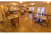 A spacious dining room with several wooden tables and chairs arranged neatly. The room has warm lighting, large windows with blinds, and a wooden floor. The walls are painted in a light color, and there is a counter area in the back with additional seating and kitchen access.