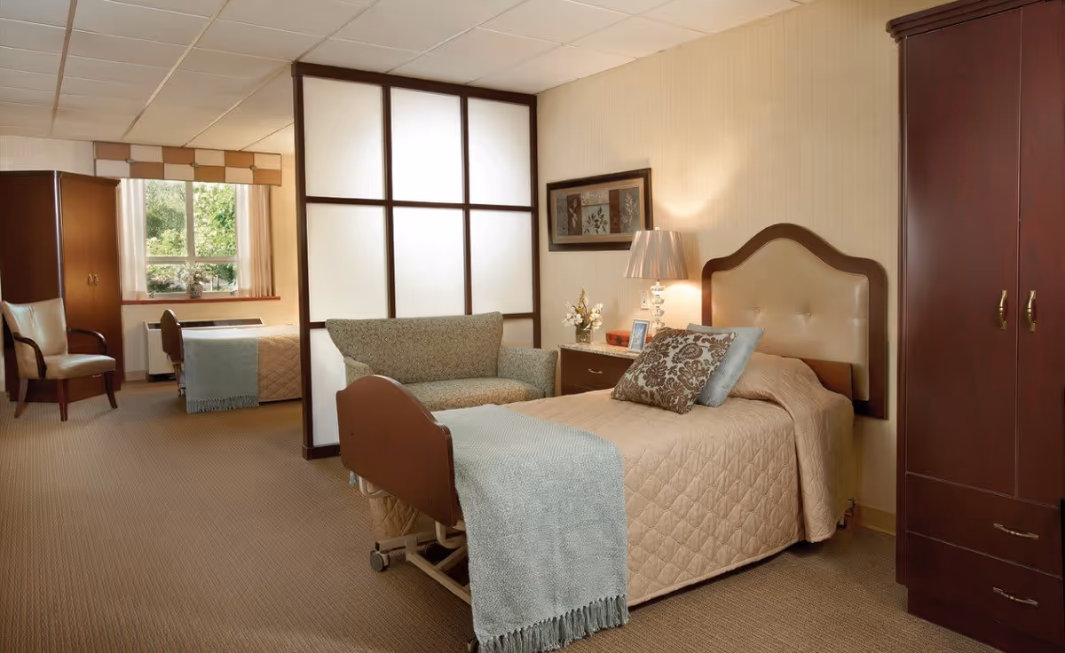 A cozy senior living bedroom featuring a single bed with a beige quilt and decorative pillows, a wooden headboard, a nightstand with a lamp and framed picture, a small sofa, a wooden wardrobe, and a window with a view of greenery. The room is softly lit and has a partition separating the sleeping area from a small sitting area with chairs and a table near the window.