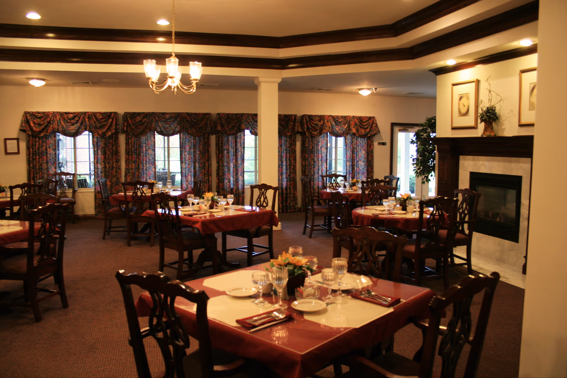 A dining room with multiple tables covered in red tablecloths and set with plates, glasses, and silverware. The room has carpeted floors, floral curtains on the windows, a chandelier hanging from the ceiling, and a fireplace with framed artwork above it.
