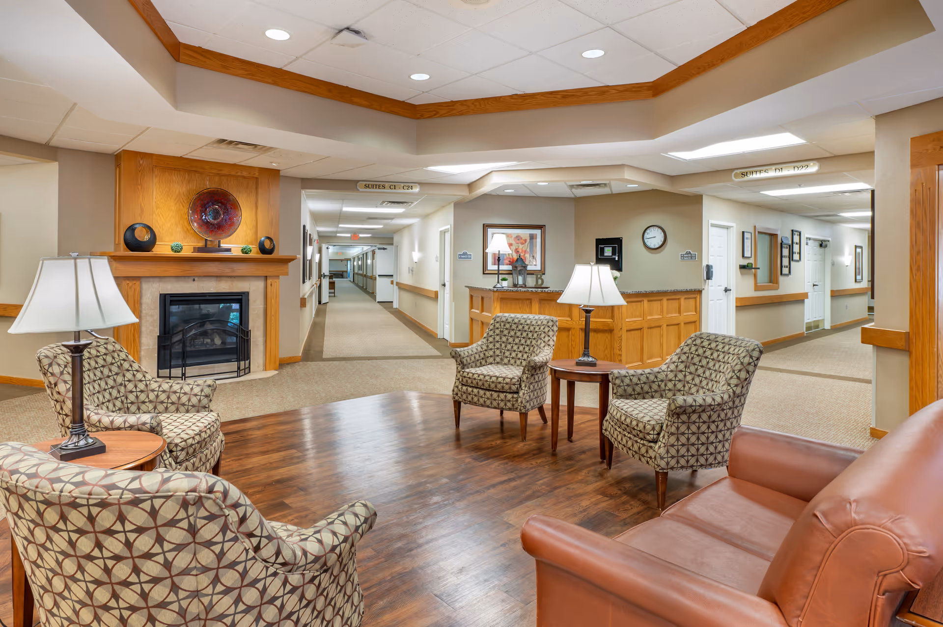 A cozy seating area in a senior living facility with four patterned armchairs and one brown leather sofa arranged around a wooden floor section. There are two table lamps on small side tables, a fireplace with decorative items above it, and a reception desk in the background. The area is well-lit with ceiling lights and has beige walls and carpeted hallways leading to different suites.