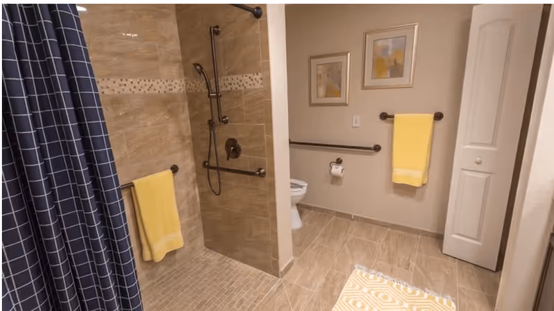 Accessible bathroom with a walk-in tiled shower, grab bars, toilet, yellow towels and a patterned rug.