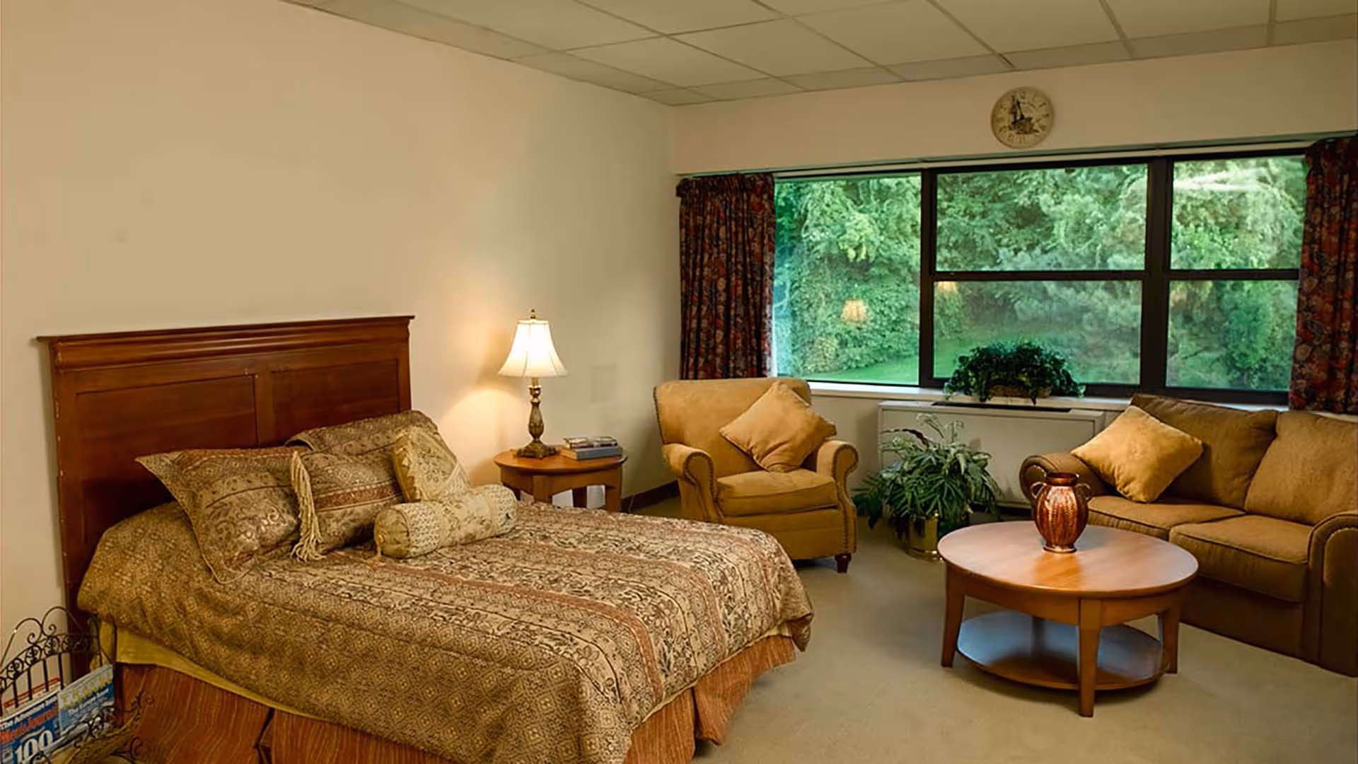 A cozy bedroom with a wooden bed frame and patterned bedding, a bedside table with a lamp and books, an armchair with a cushion, a sofa with cushions, a round wooden coffee table with a decorative vase, large window with floral curtains showing green trees outside, and indoor plants near the window.