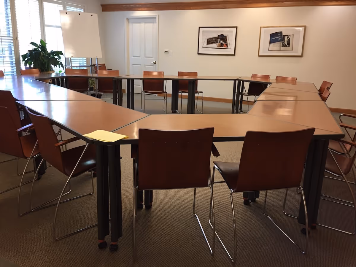 Conference-style activity room with U-shaped wooden tables and brown chairs, a whiteboard, plant, and framed pictures on the wall.
