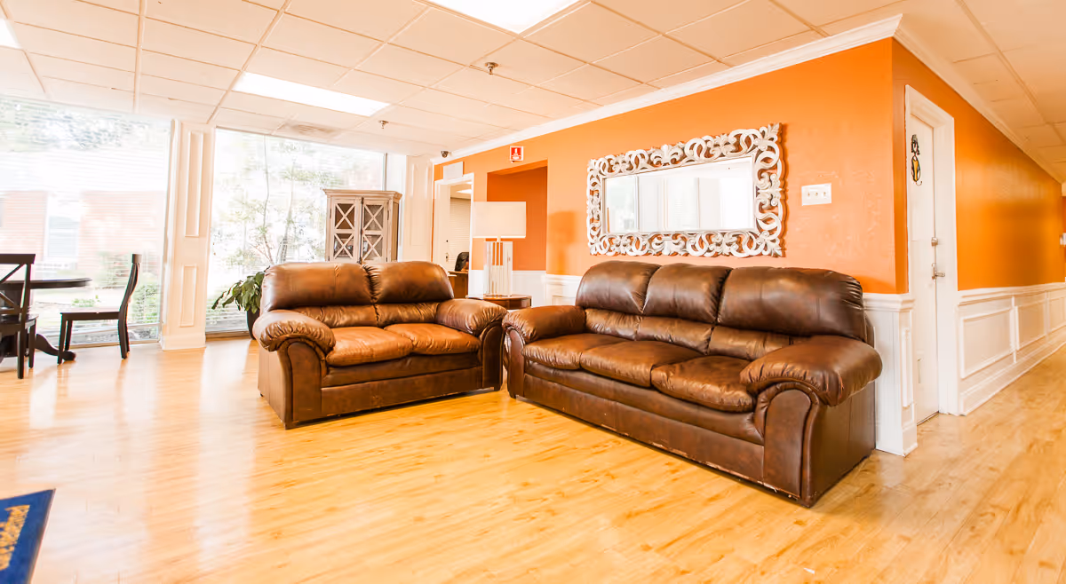 A bright and spacious living room area with two brown leather sofas placed against an orange wall. A decorative white-framed mirror hangs above the sofas. The room has light wood flooring, a large window letting in natural light, and a small table with chairs visible in the background.