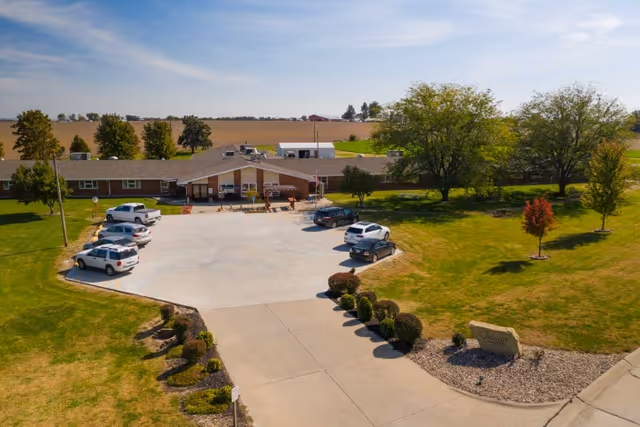 Aerial view of Falls City Nursing and Rehabilitation Center showing a single-story building with a large parking lot in front. Several cars are parked, and the surrounding area includes green lawns, trees, and open fields under a partly cloudy sky.